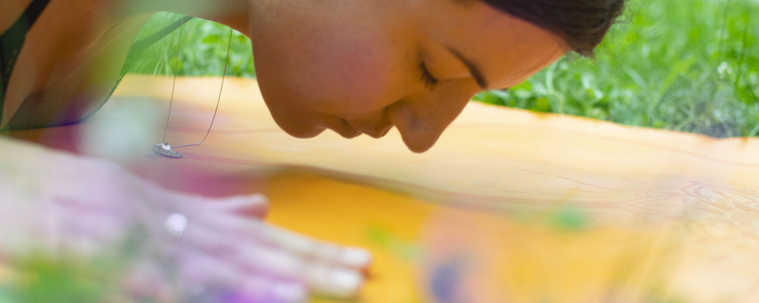 Eine Frau praktiziert Yoga auf einer Yogamatte in einer natürlichen Umgebung. Neben ihr stehen Chakra Flower Yogamattenreiniger und weitere Reinigungsprodukte. Das Bild vermittelt die Verbundenheit zwischen Yoga, natürlicher Reinigung und der umliegenden Natur